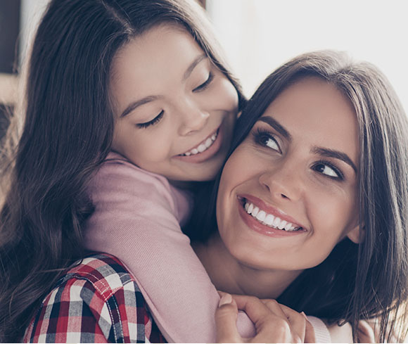 a girl hugging her mom while both are smiling showing their perfect teeth