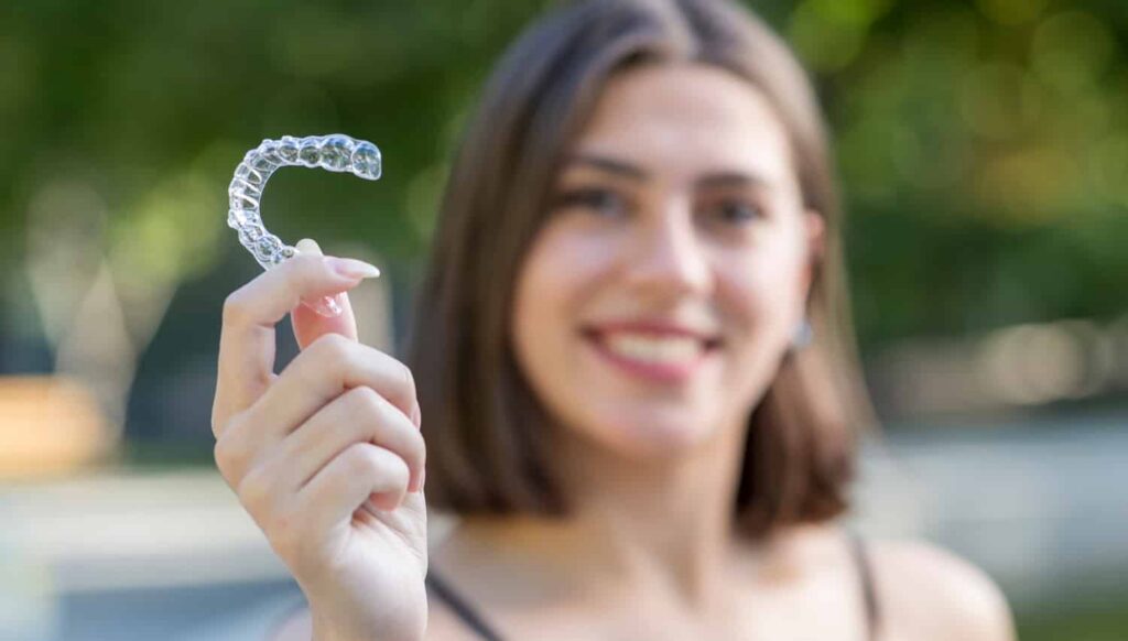 Young woman smiling while holding a clear Invisalign aligner, showcasing its use for orthodontic treatment.