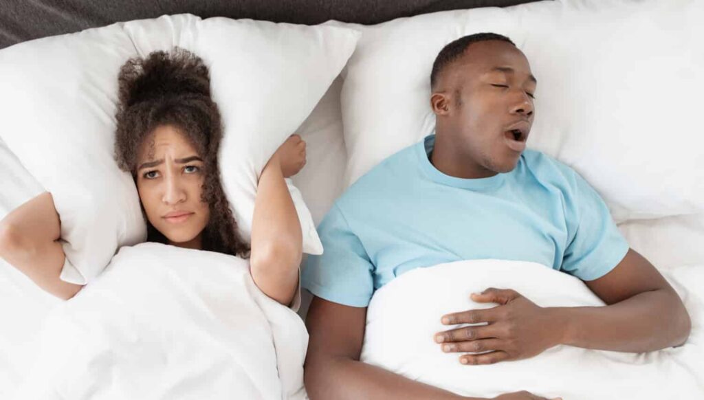 Couple in bed, woman covering ears, looking distressed, man snoring, illustrating the impact of snoring and sleep apnea on relationships.