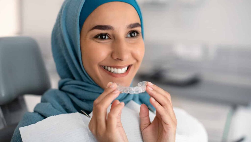 Smiling woman wearing a hijab holding Invisalign clear aligners in a dental office setting, representing modern teeth straightening options.