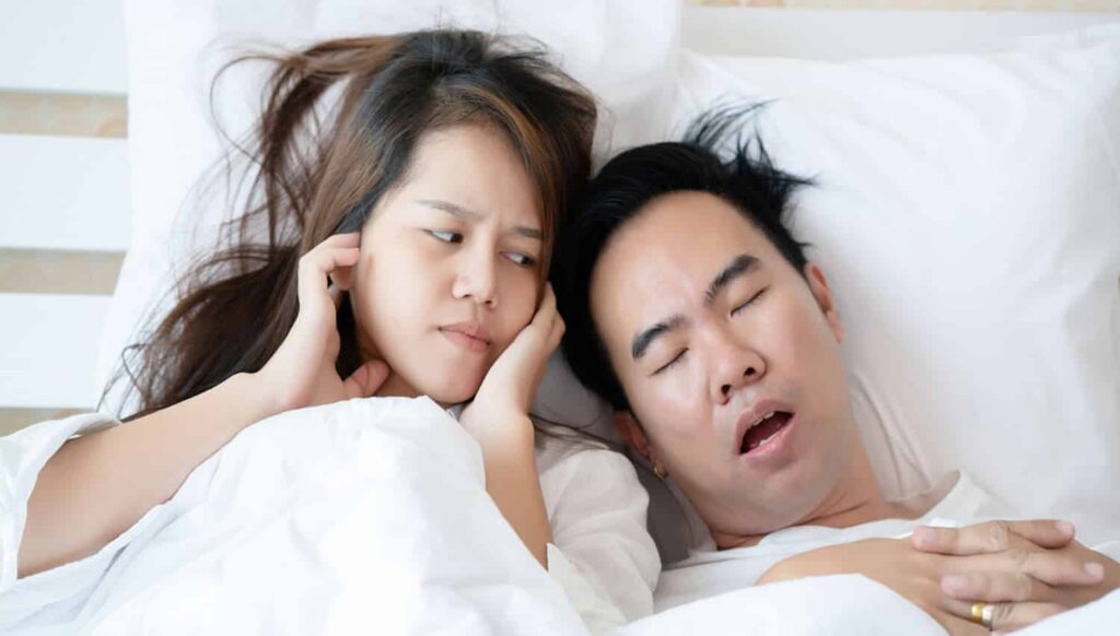 Couple in bed, woman covering ears with a frustrated expression, man snoring, illustrating sleep apnea's impact on partners.