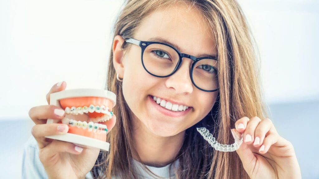 Girl smiling while holding a model of braces and an Invisalign aligner, illustrating the comparison between orthodontic treatments.