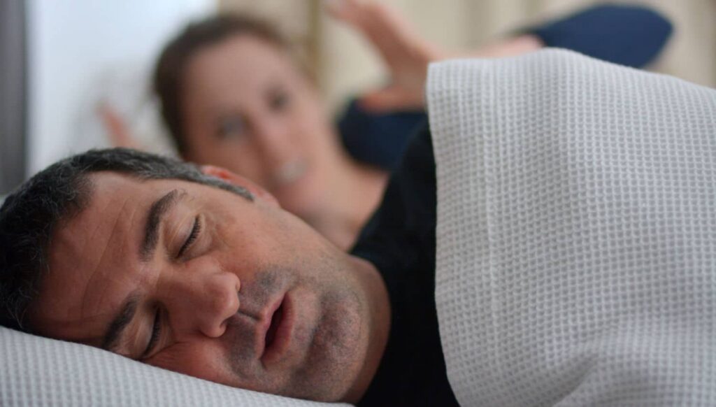 Man sleeping soundly while a woman in the background appears frustrated, illustrating the impact of snoring on partners.