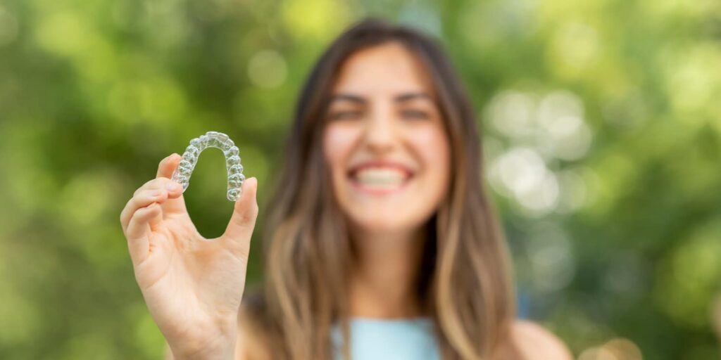 Smiling woman holding clear Invisalign aligner outdoors, showcasing benefits of invisible orthodontics.