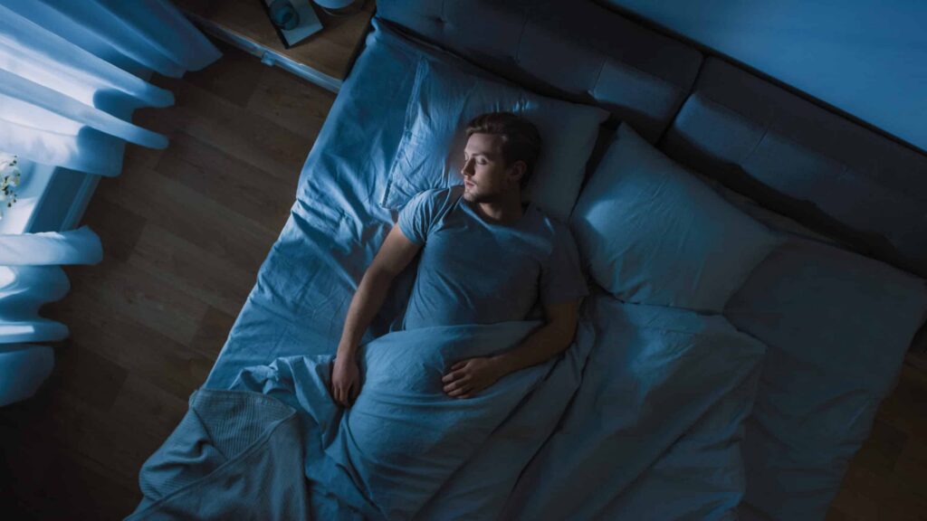 Man lying awake in bed, surrounded by blue bedding and pillows, illustrating the struggles of sleep apnea and the importance of quality sleep in relation to treatment options like the Vivos System.