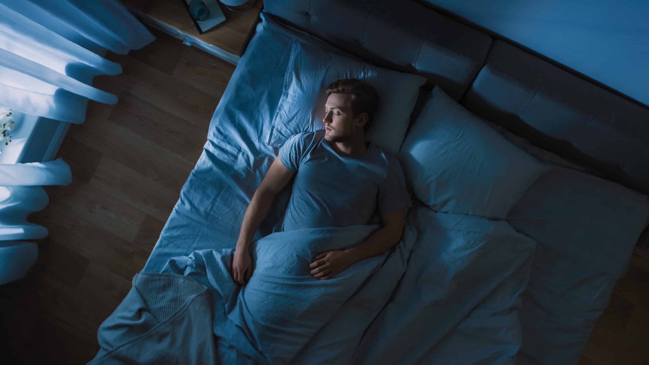 Man lying in bed, appearing to sleep peacefully, with soft blue lighting and curtains, illustrating the theme of sleep health in the context of sleep apnea discussions.
