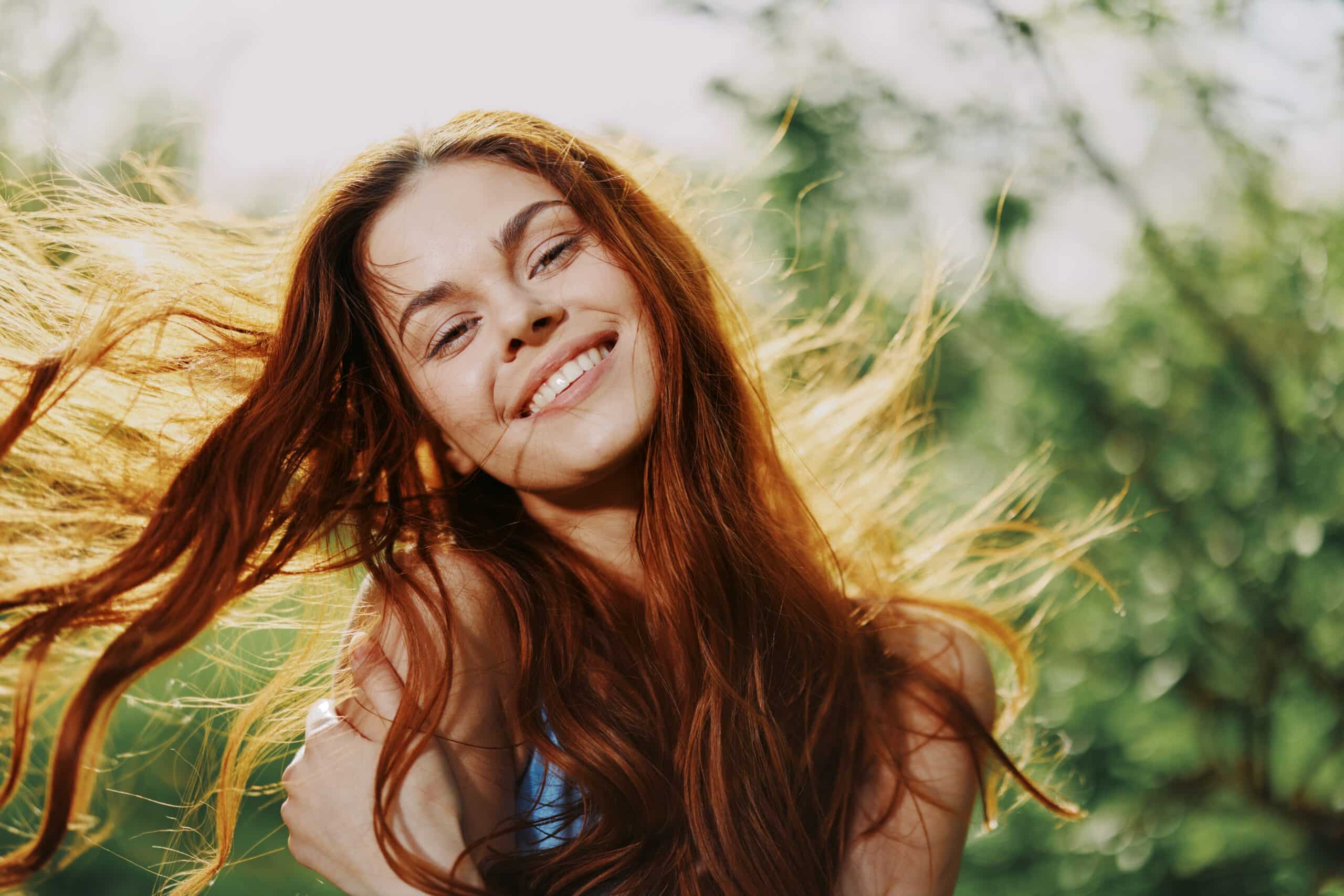 Smiling woman with long, flowing hair in a natural outdoor setting, reflecting confidence and vitality, relevant to dental health and the importance of a vibrant smile.