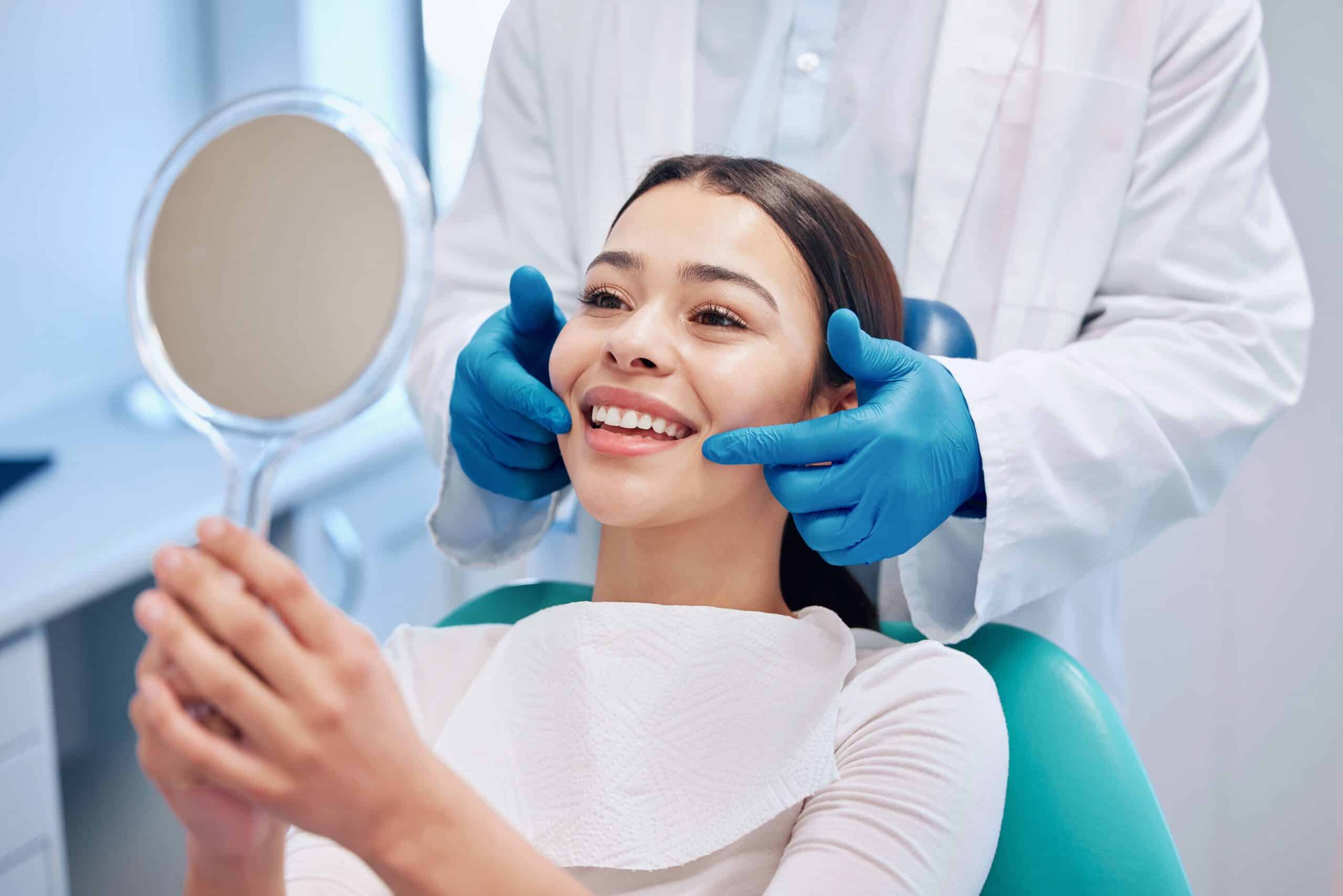 Woman smiling in dental chair, holding mirror, dentist in gloves examining her teeth, dental care setting.