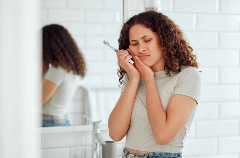 Woman experiencing toothache while holding her face, in a bathroom setting, illustrating common dental pain related to tooth decay and sensitivity.