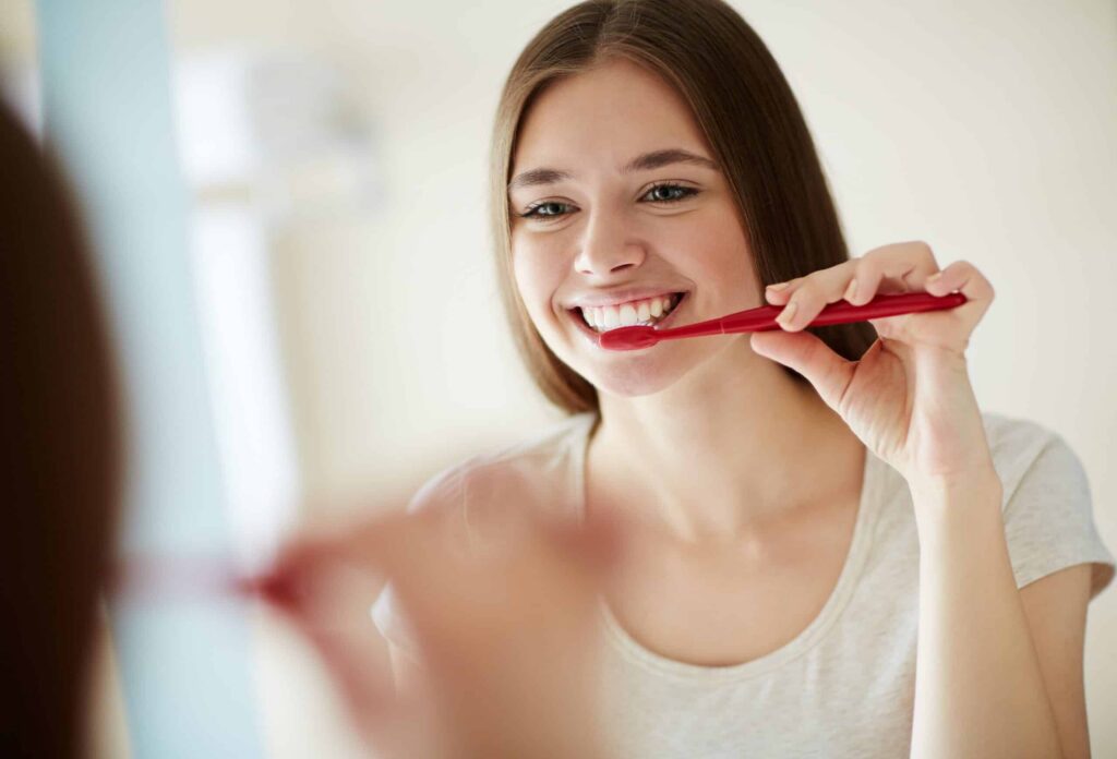 Woman brushing her teeth with a red toothbrush, smiling in front of a mirror, promoting good oral hygiene for healthy gums and a bright smile.