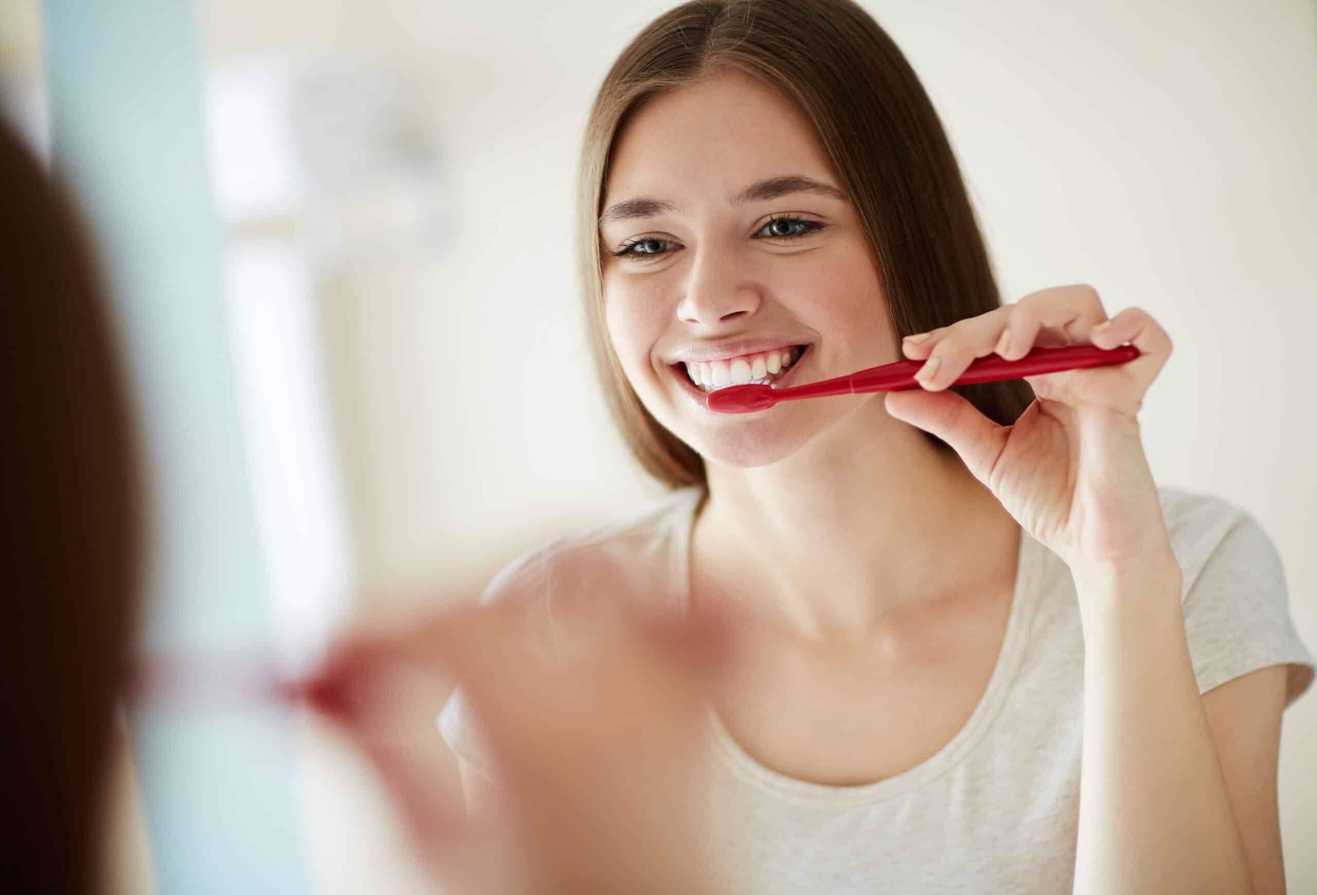 Young woman brushing her teeth with a red toothbrush, smiling in front of a mirror, promoting healthy gums and oral hygiene.