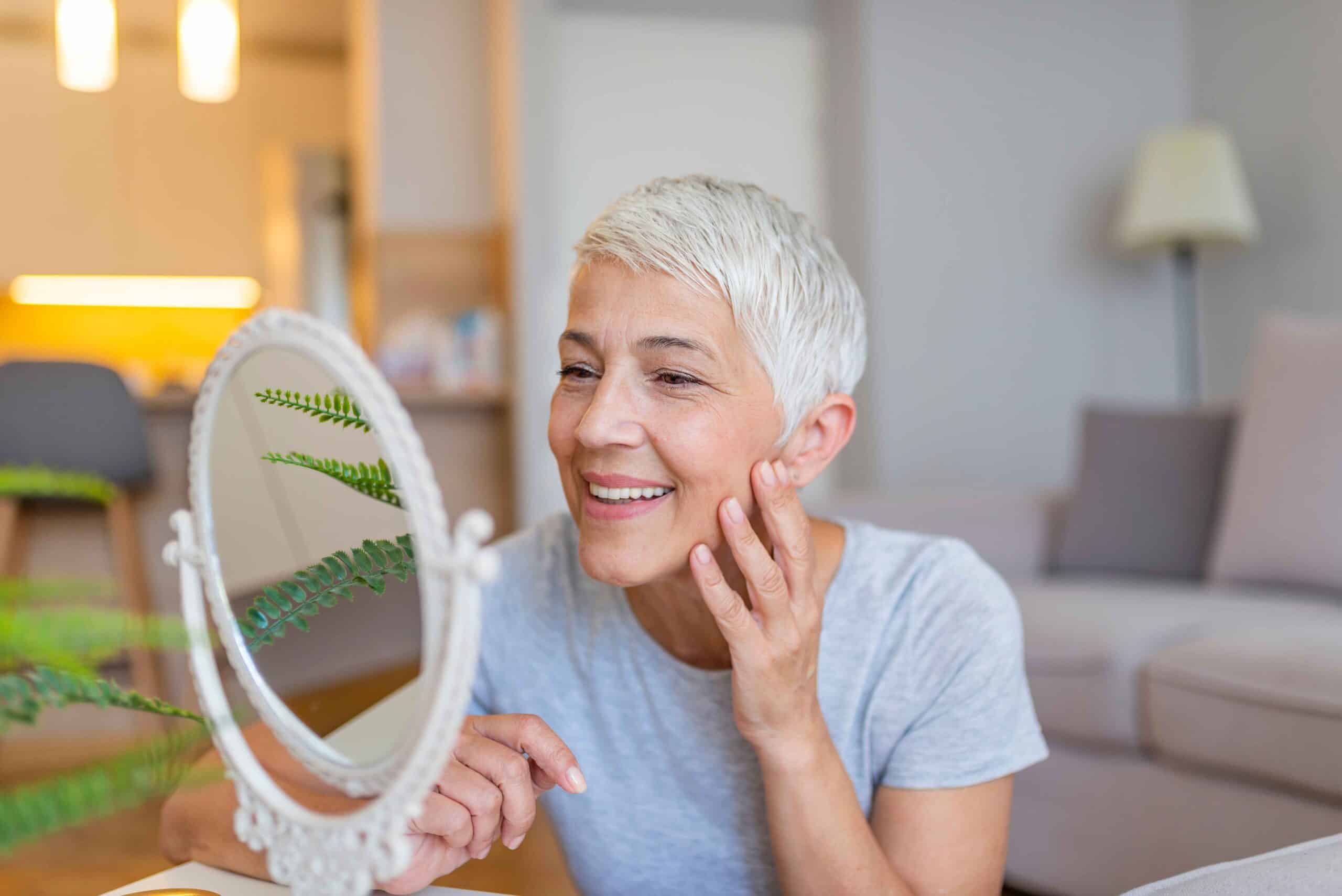Smiling older woman examining her reflection in a mirror, emphasizing the importance of dental care and oral health for the aging population.