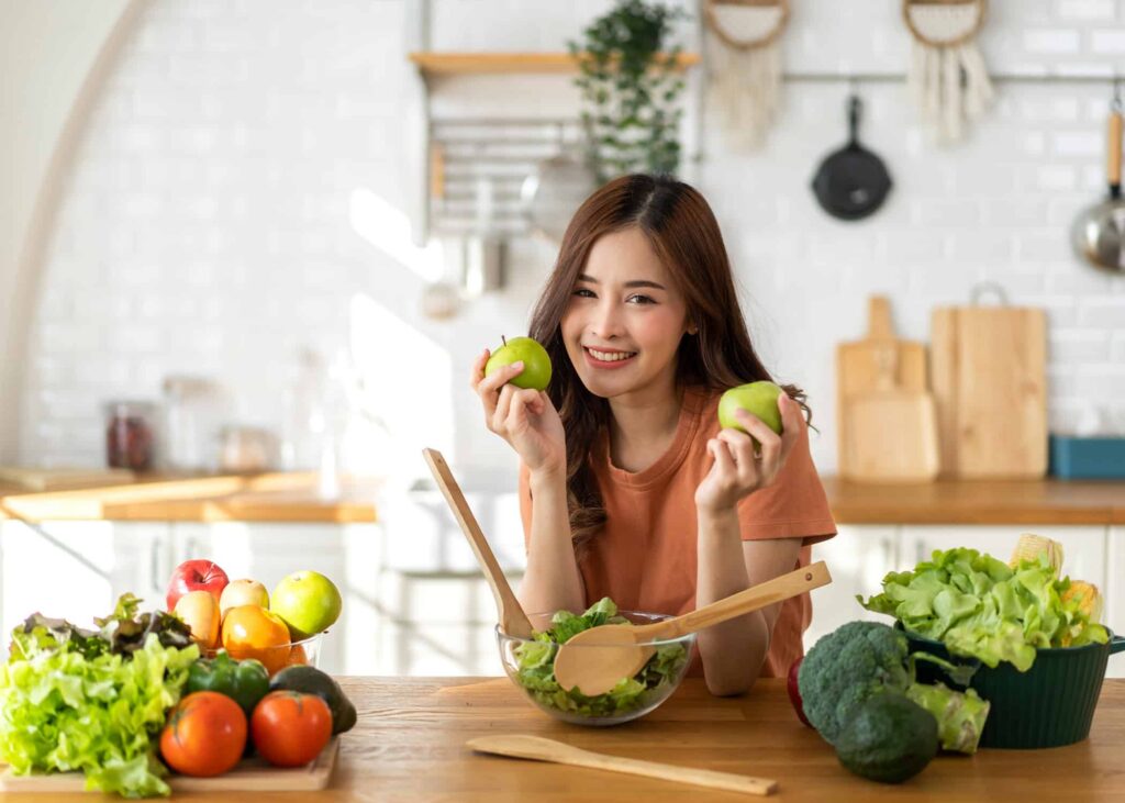 Woman holding green apples and smiling, surrounded by fresh vegetables and salad ingredients, emphasizing nutrition's role in promoting dental health for a healthy smile.