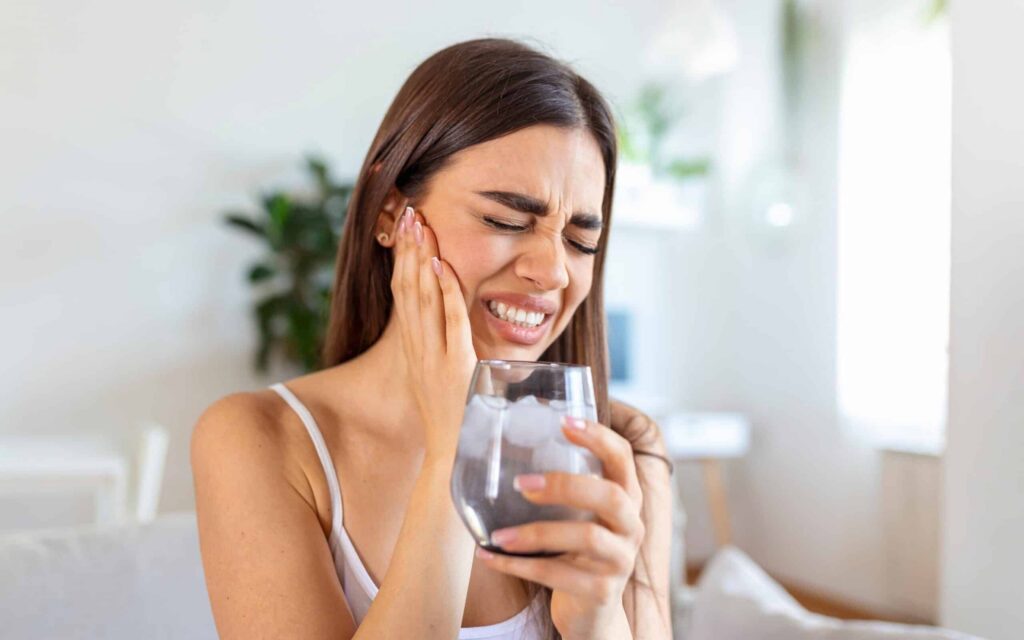 Woman experiencing tooth sensitivity while drinking cold water, holding her cheek in discomfort.