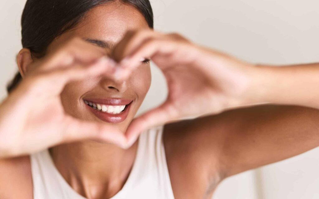 Smiling woman forming a heart shape with her hands, showcasing bright, white teeth, emphasizing the positive effects of teeth whitening.