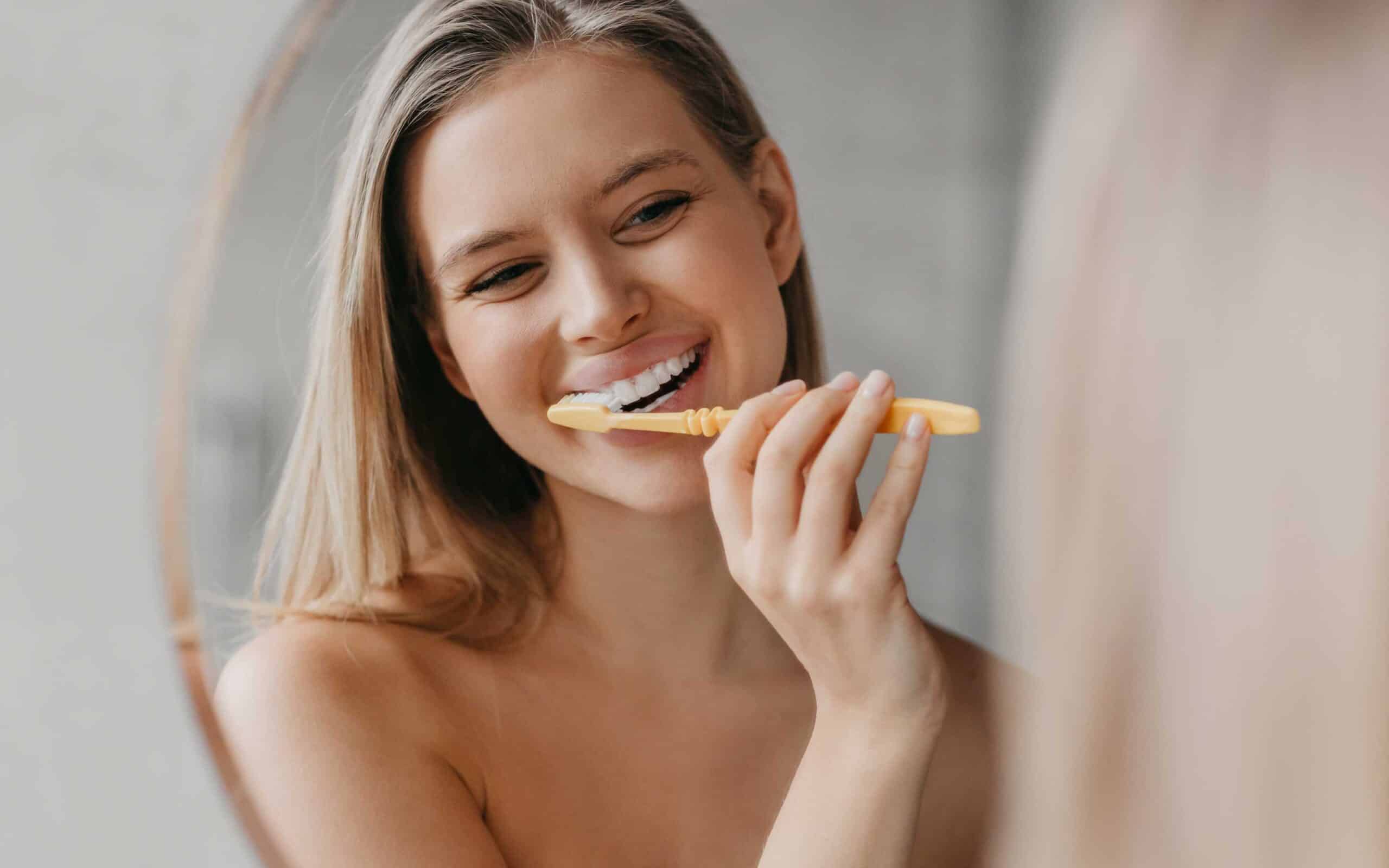 Young woman brushing her teeth with a yellow toothbrush in front of a mirror, emphasizing daily dental hygiene and preventive care.