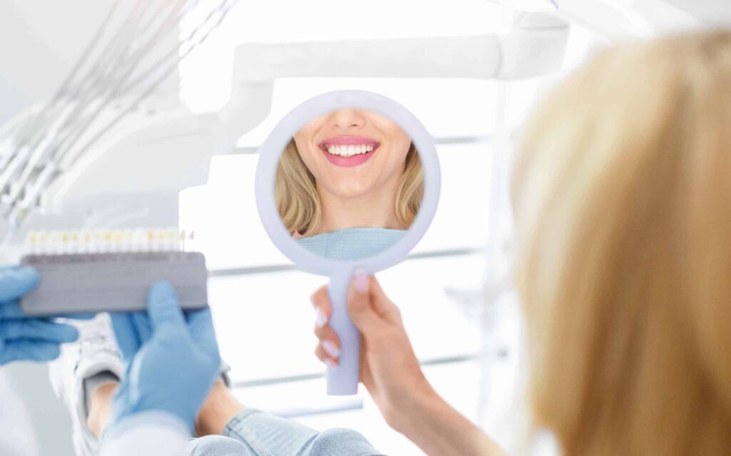 Woman smiling while looking in a dental mirror, with a dental professional holding a shade guide, illustrating the importance of dental care and coverage options at Smith Dentalworks in Oro Valley, Arizona.