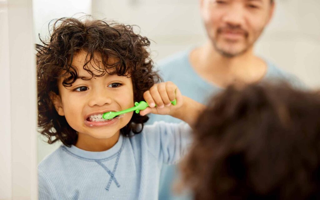 Child brushing teeth with green toothbrush, emphasizing early dental care and healthy habits, with parent observing in background.