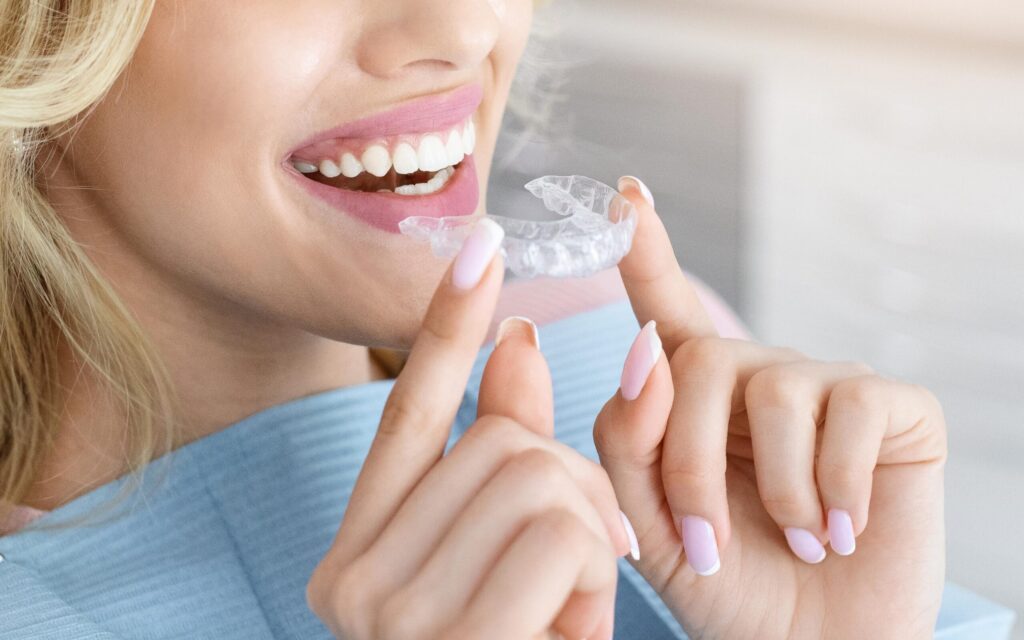 Woman holding Invisalign clear aligner near her mouth, showcasing a bright smile, illustrating modern orthodontic solutions for adults considering Invisalign treatment.