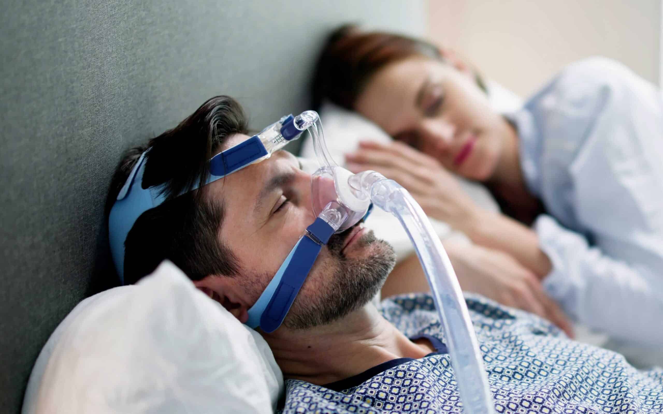 Man using a CPAP machine for sleep apnea treatment while a woman sleeps beside him, highlighting the connection between sleep apnea and oral health.