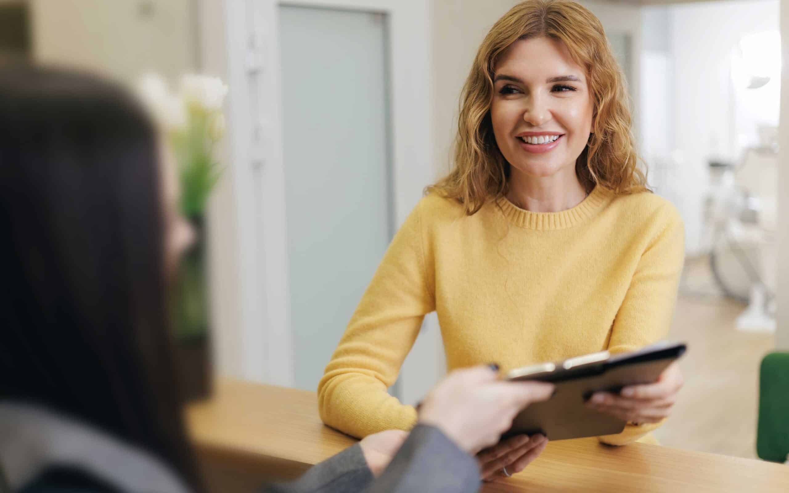 Smiling woman in a yellow sweater discussing dental services at a reception desk, emphasizing patient care and accessibility at a full-service dental office.