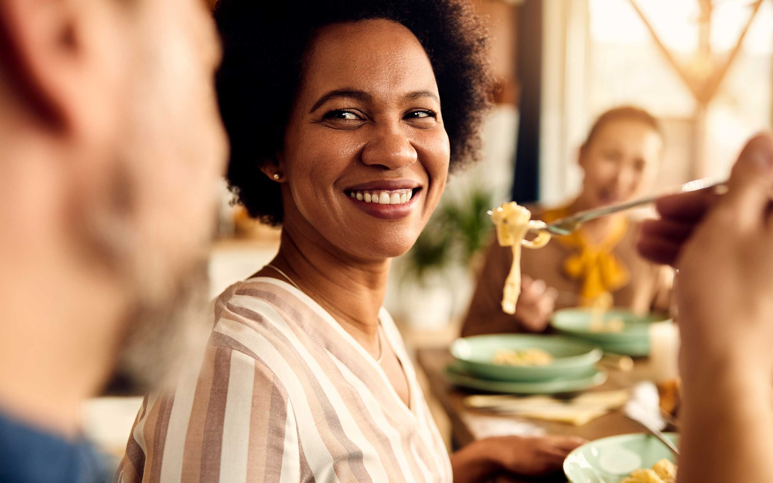 Smiling woman enjoying a meal with pasta, emphasizing the connection between diet and oral health in a social setting.