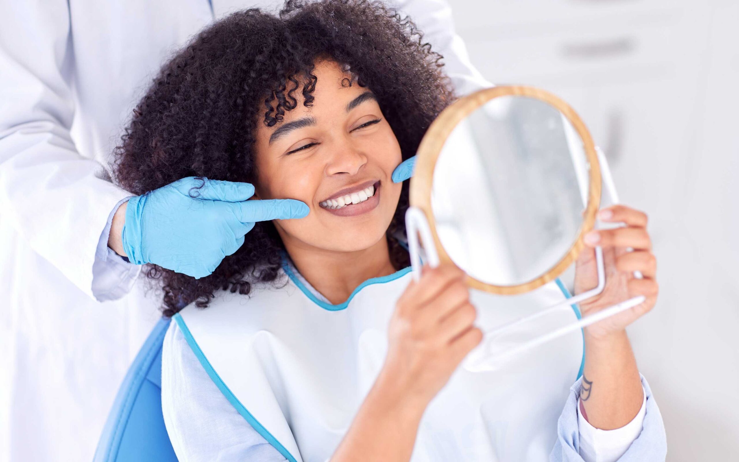 Woman smiling while examining her teeth in a mirror after a teeth whitening treatment, illustrating teeth whitening cost and cosmetic dental care.